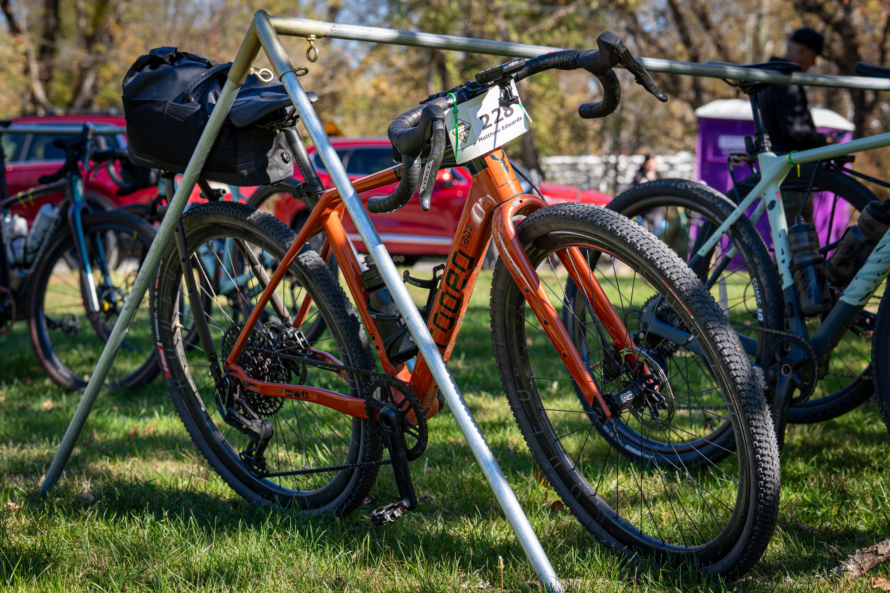 A burnt-orange bike is resting on a bike rack, surrounded by other bikes. The scene is set on grassy ground under clear blue skies.